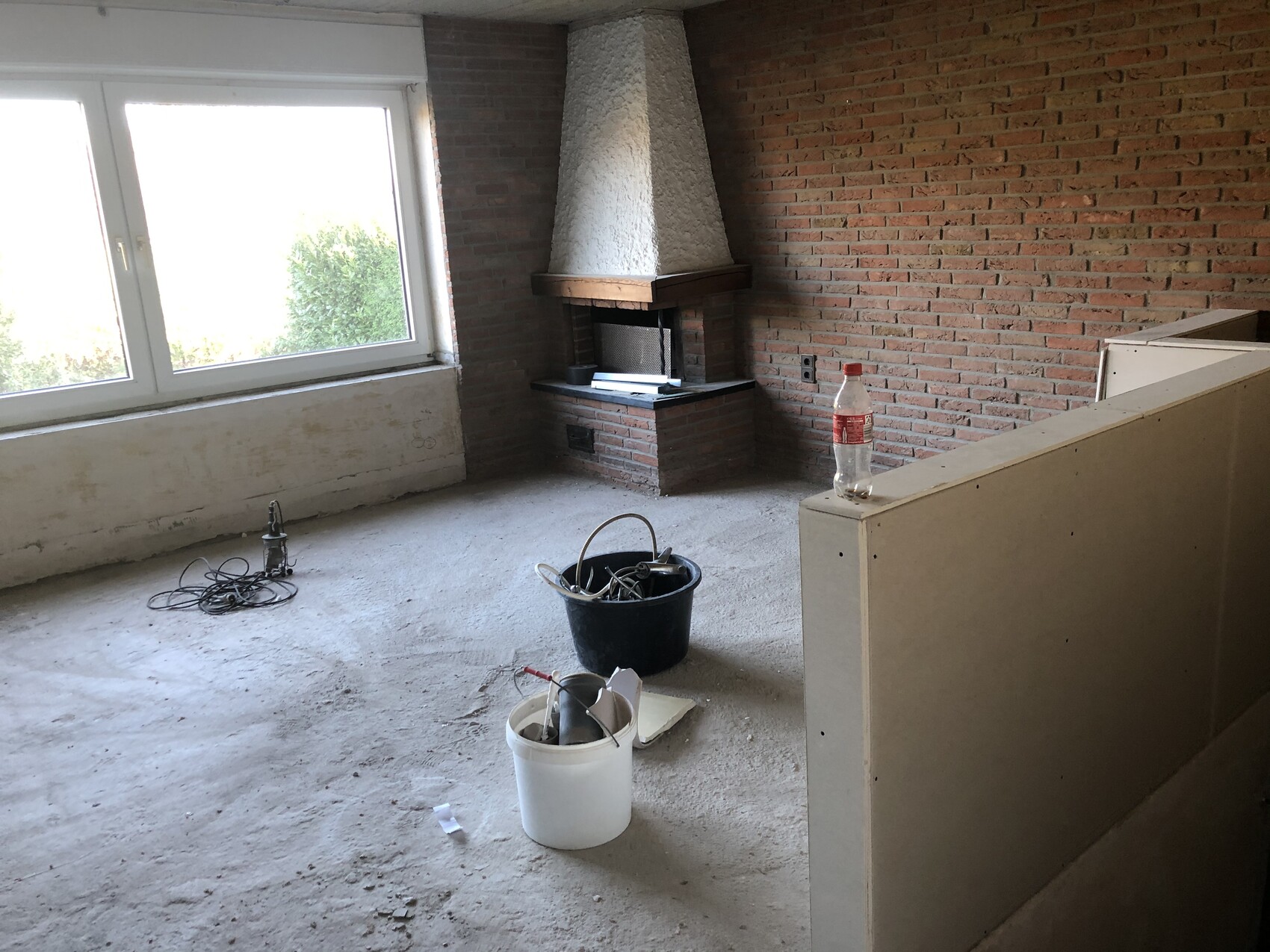 Unrenovierter Raum mit Kamin, Ziegelwand, großem Fenster links, Eimer, Kabeln und Flasche.