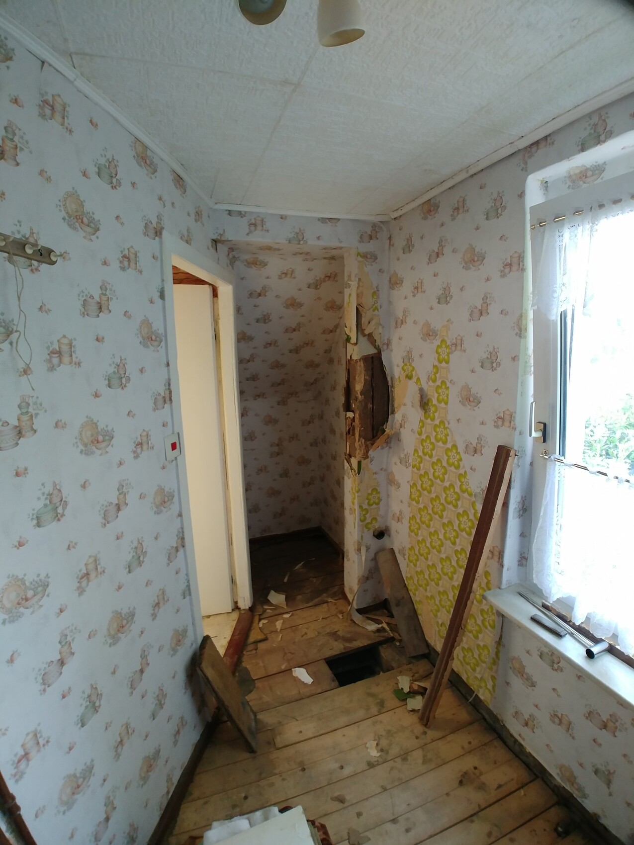 Ein renovierungsbeduerftiger Flur mit beschädigter Wandverkleidung, Tapete abgerissen und Loch im Boden