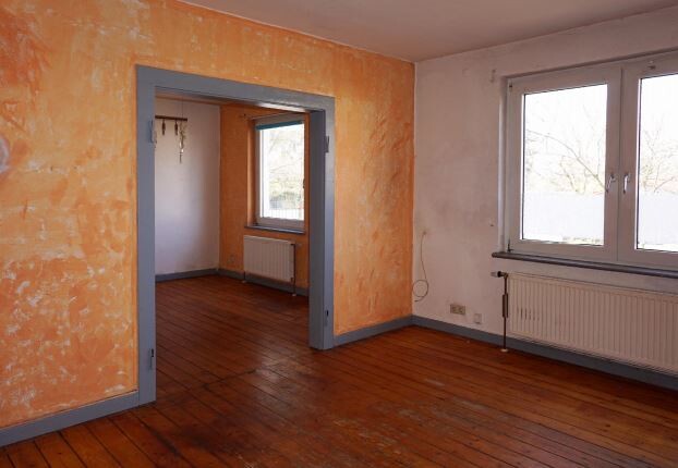 Wohnzimmer mit orange verputzter Wand links, Durchgang zu anderem Raum, Holzboden, Fenster rechts.