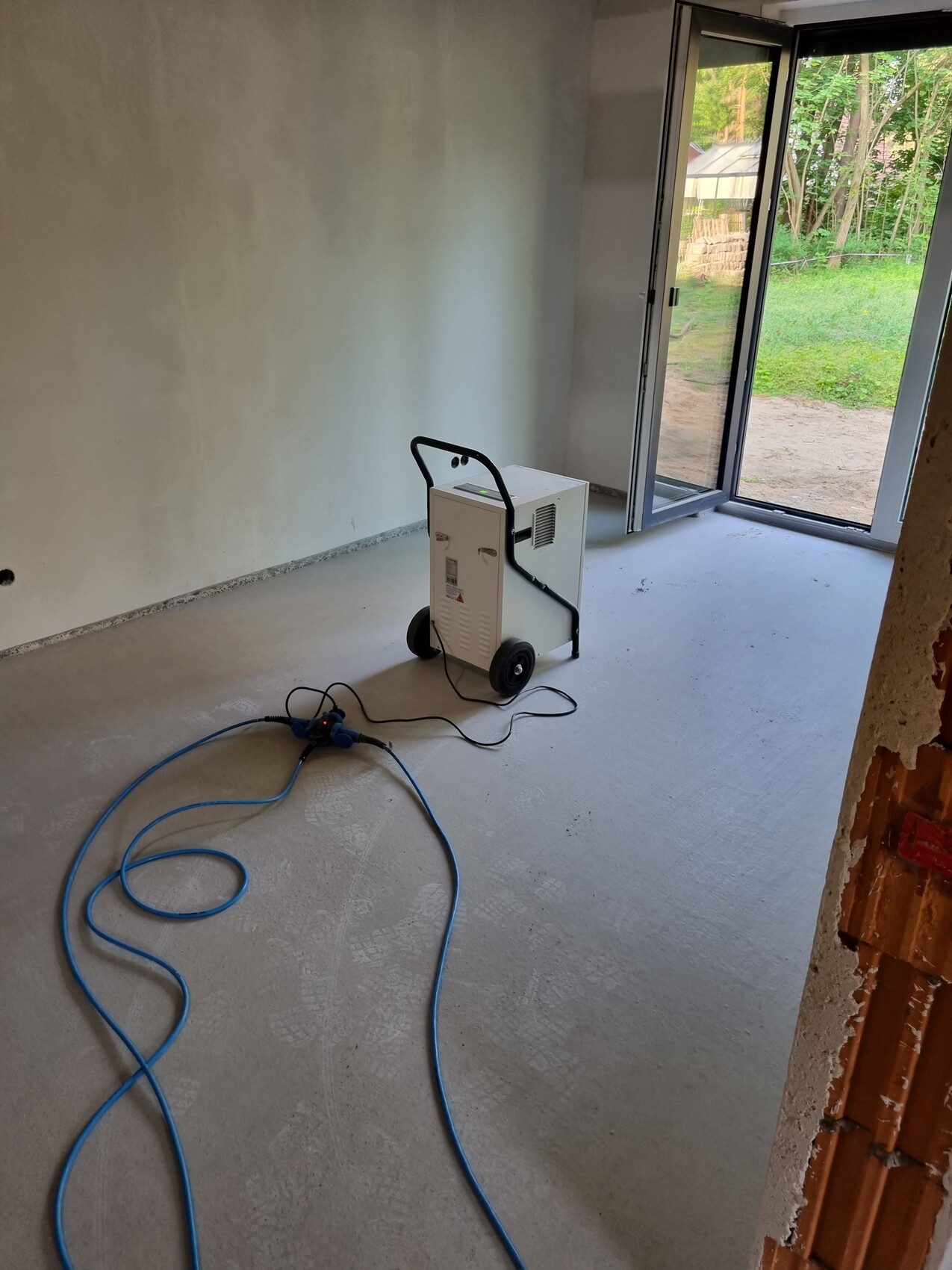 Mobiler Entfeuchter auf Rädern mit blauem Schlauch im unfertigen Zimmer, Terrassentür offen.