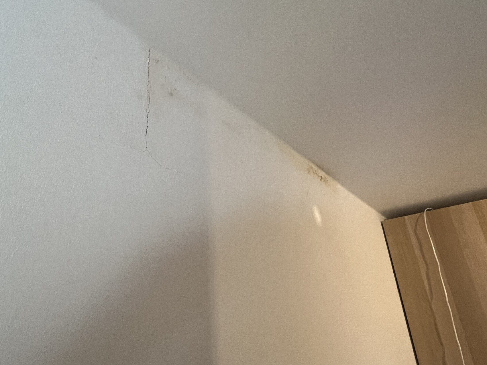 Ecke eines Zimmers: Wandrisse an der linken Wand, diagonaler Lichtstreifen, Holzschrank mit Kabel.