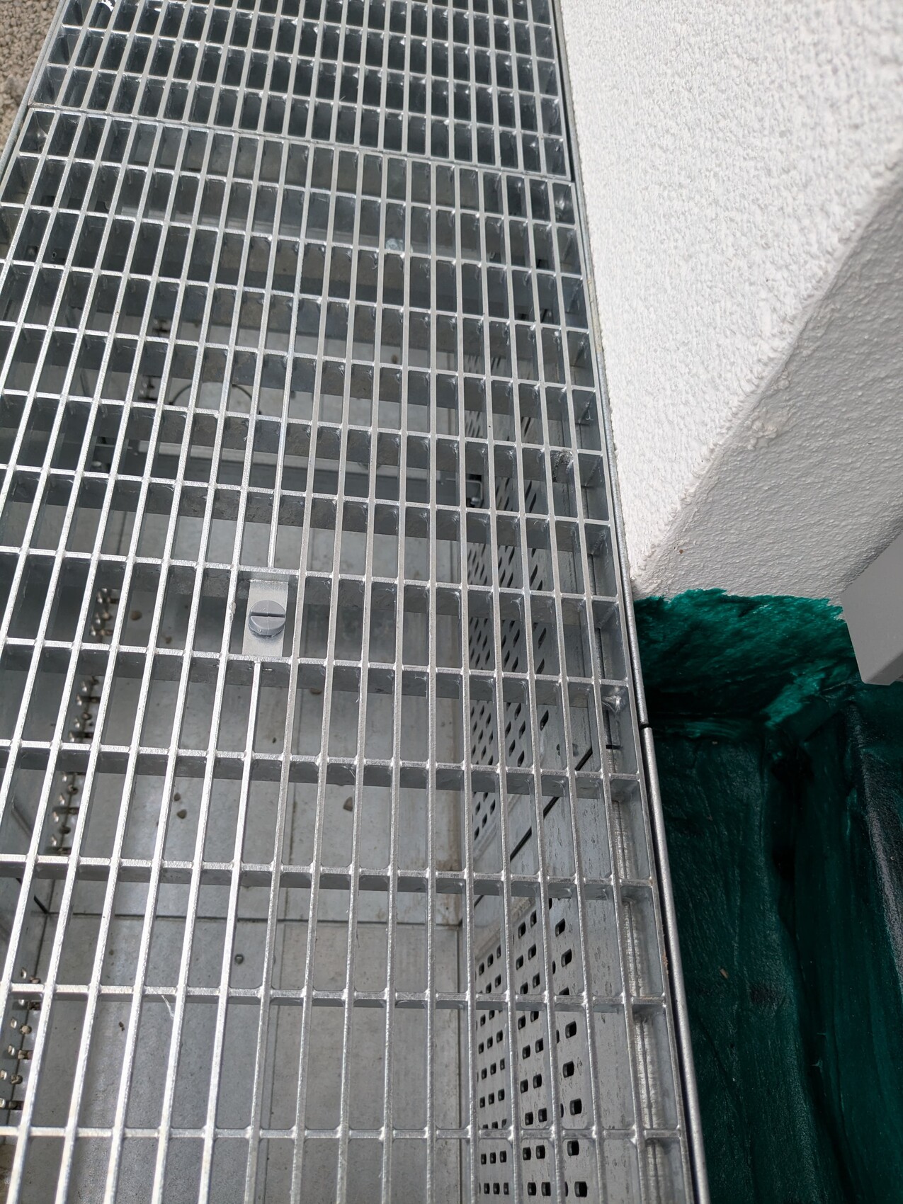 Metallgitterboden über einem offenen Schacht; graue Innenfläche, grüne Plane neben weißer Wand.