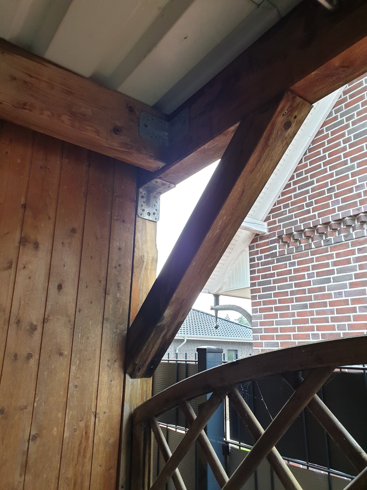 Holzbalken-System mit diagonalem Balken und Metallwinkeln; Backsteinwand im Hintergrund.