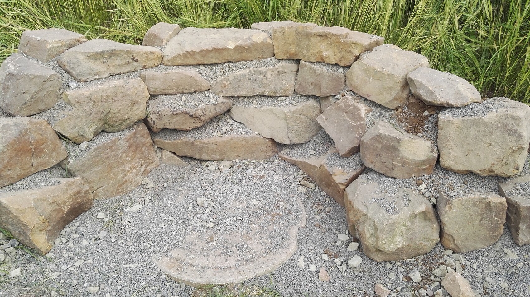 Natursteinmauer aus groben Steinen, Kiesboden darunter, hohe Gräser dahinter.