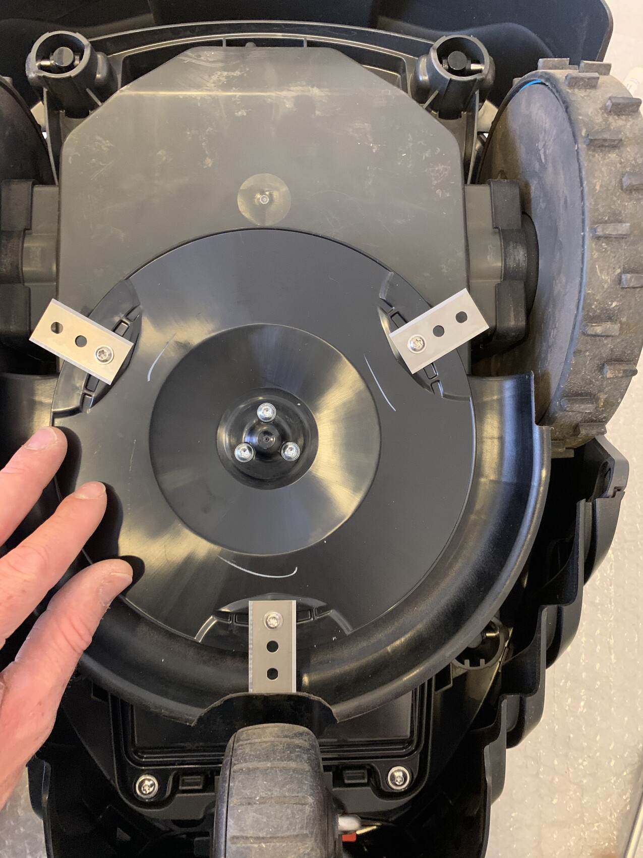 Nahaufnahme eines Rasenmähers: runde schwarze Abdeckung, Metallhalterungen mit Löchern, großes Rad sichtbar.
