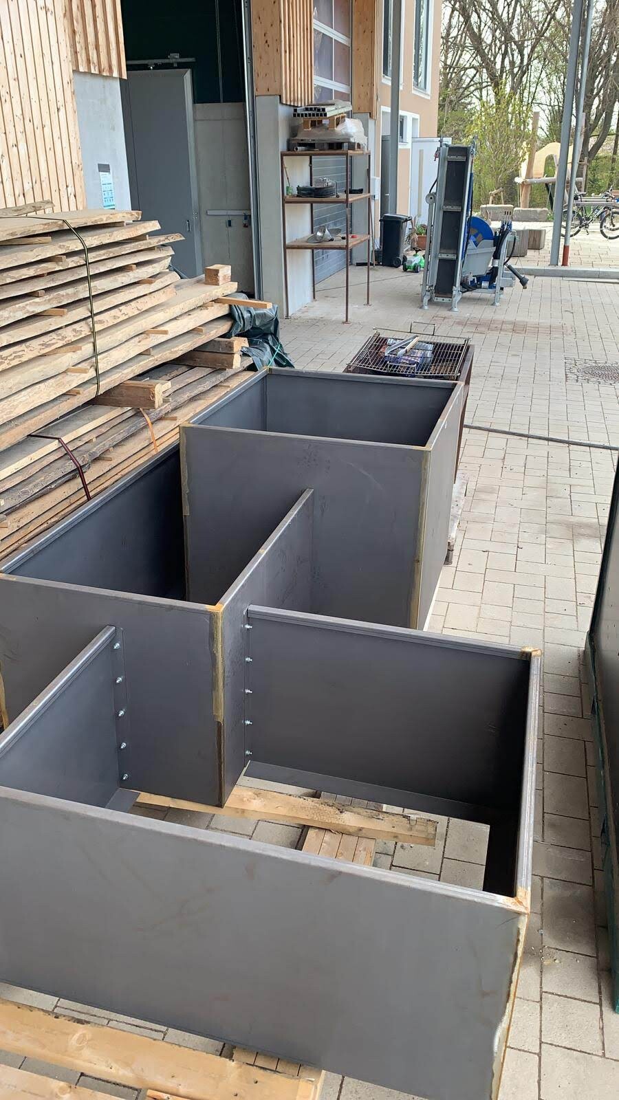 Mehrere große graue Metallbehälter stehen im Außenbereich einer Werkstatt, neben Holzplankenstapeln.