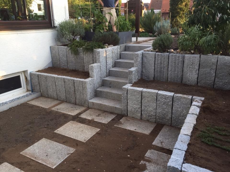 Garten mit Stützmauer aus Betonblöcken, Treppe und Pflanzbeeten.