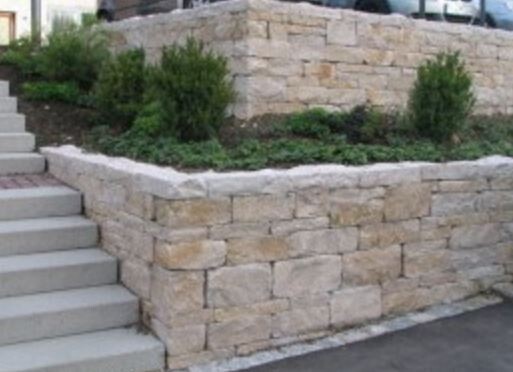 Stützmauer aus hellen Natursteinblöcken mit linksseitiger Treppe und bepflanztem Gartenbereich.