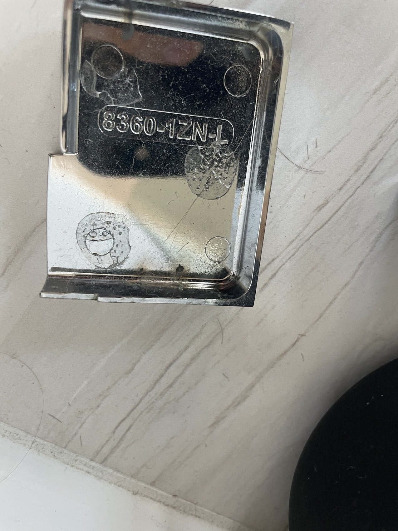 Nahaufnahme einer glänzenden Metall-Halterung mit der Gravur 8360-1ZN-T auf hellem Untergrund.
