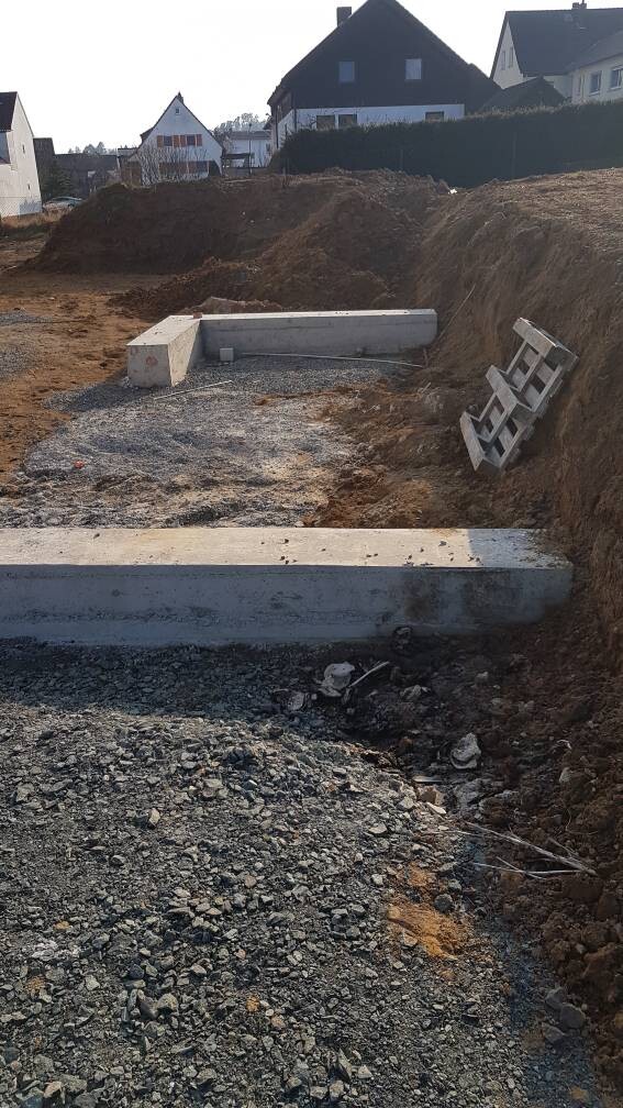 Baugrube mit Betonfundament, Kies und Erdhügeln; Häuser im Hintergrund.