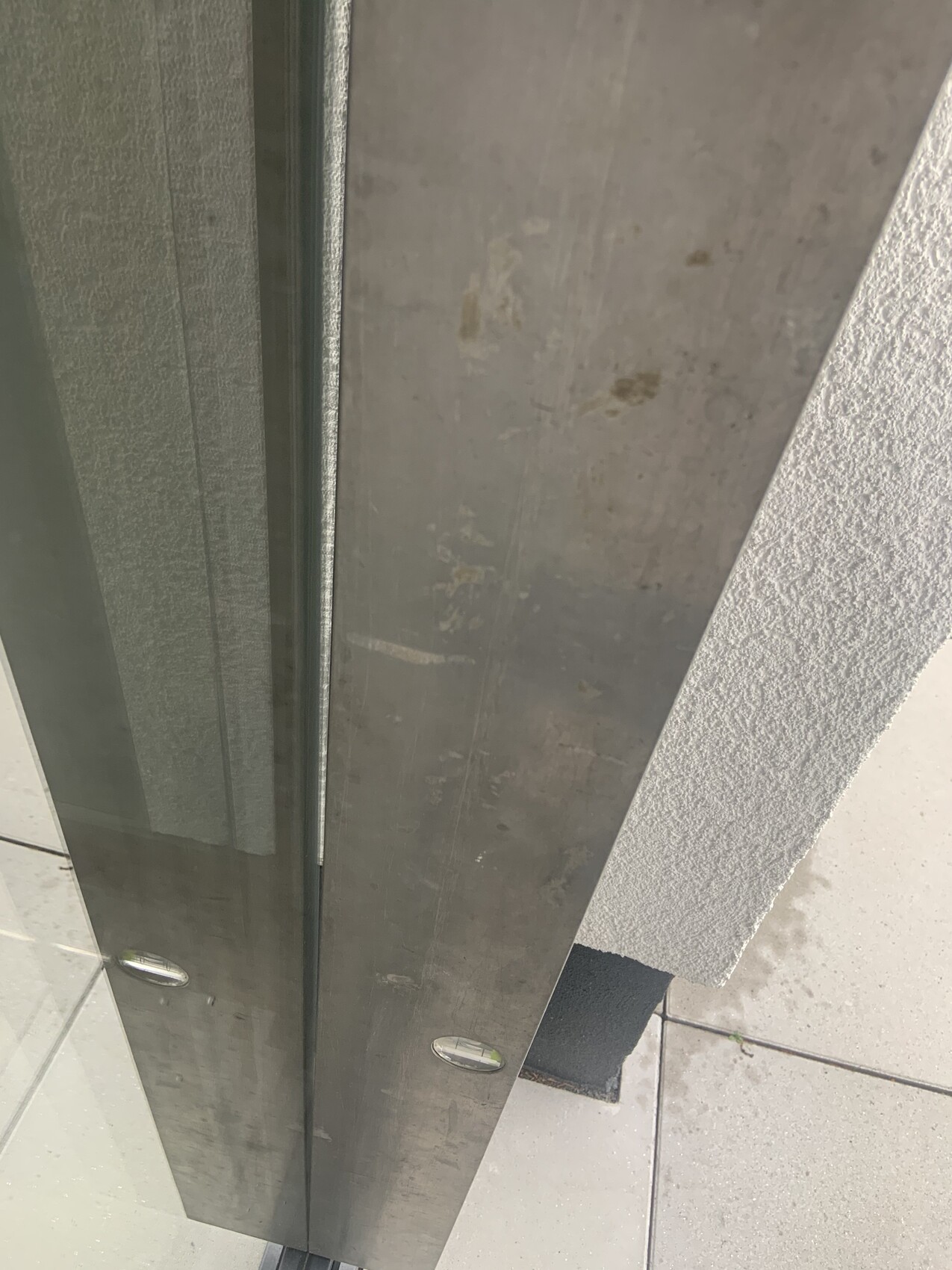Nahaufnahme eines Edelstahl-Türrahmens mit Glas und rauer weißer Wand daneben.