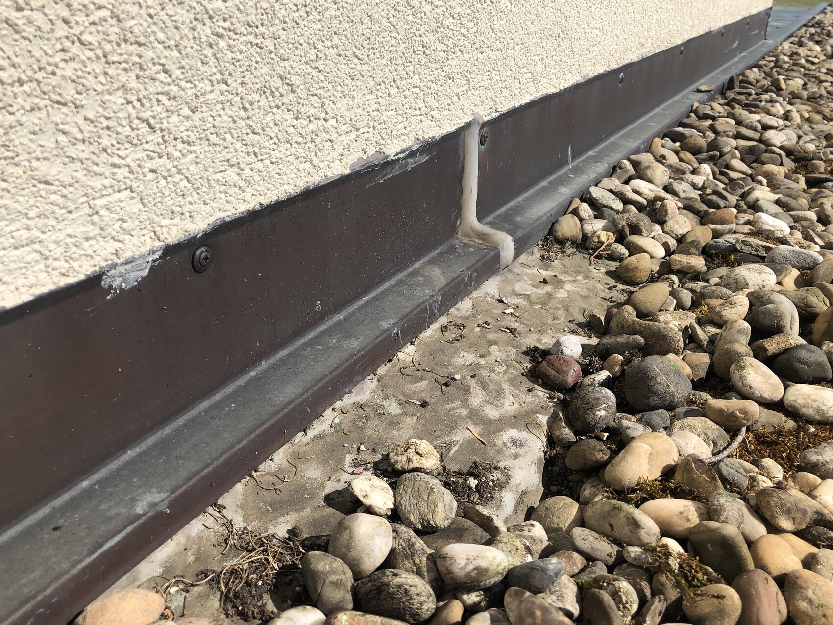 Außenwand mit brauner Sockelverkleidung, Dichtungsmasse und Kieselsteine am Fundament.