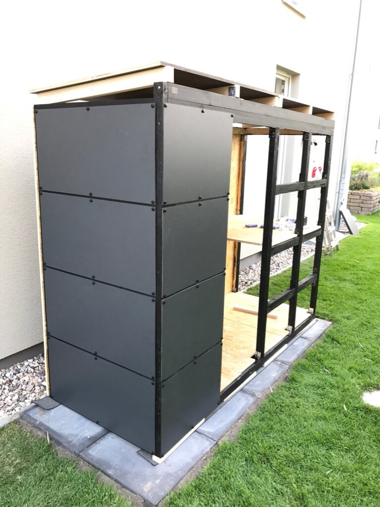 Offene Gartenkonstruktion aus schwarzem Metallrahmen mit grauen Wandplatten vor Haus.