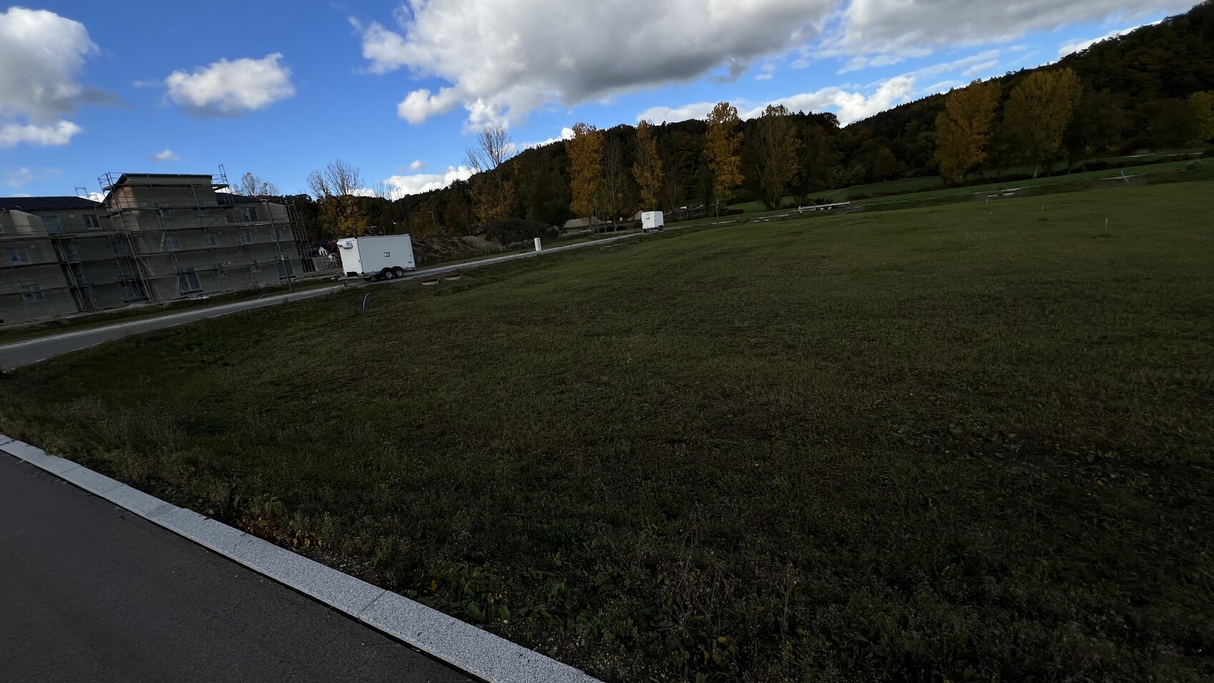 Rohbau-Baustelle mit Gerüst, Bauwagen und offener Gruenflaeche vor Baumreihe
