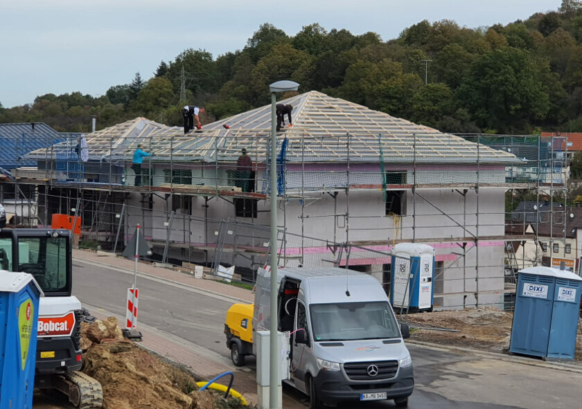 Bauarbeiter montieren Dachstuhl eines Neubaus auf Gerüst; Baumaschinen und Porta-Potties sichtbar