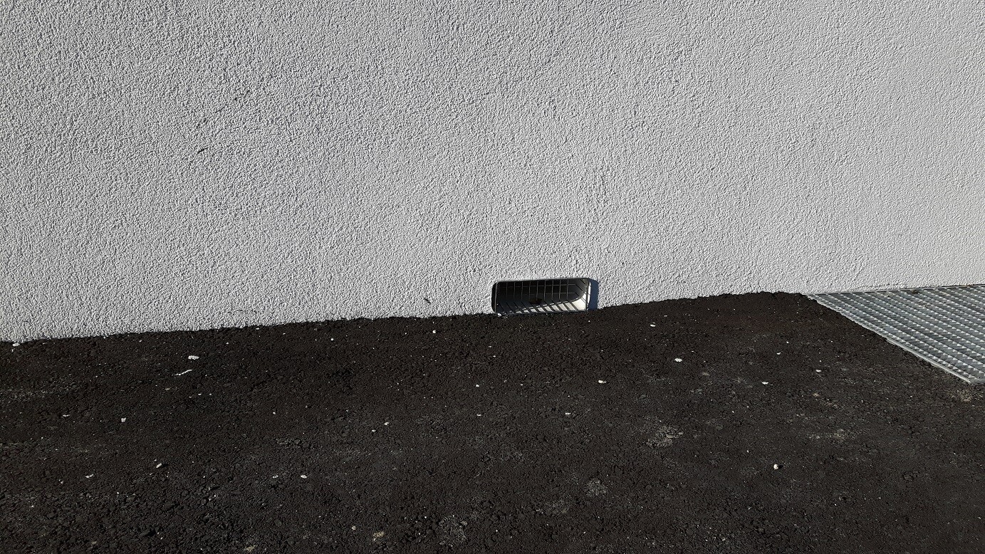 Außenwand mit rauem Putz, dunkles Erdreich und Metallbodenabdeckung
