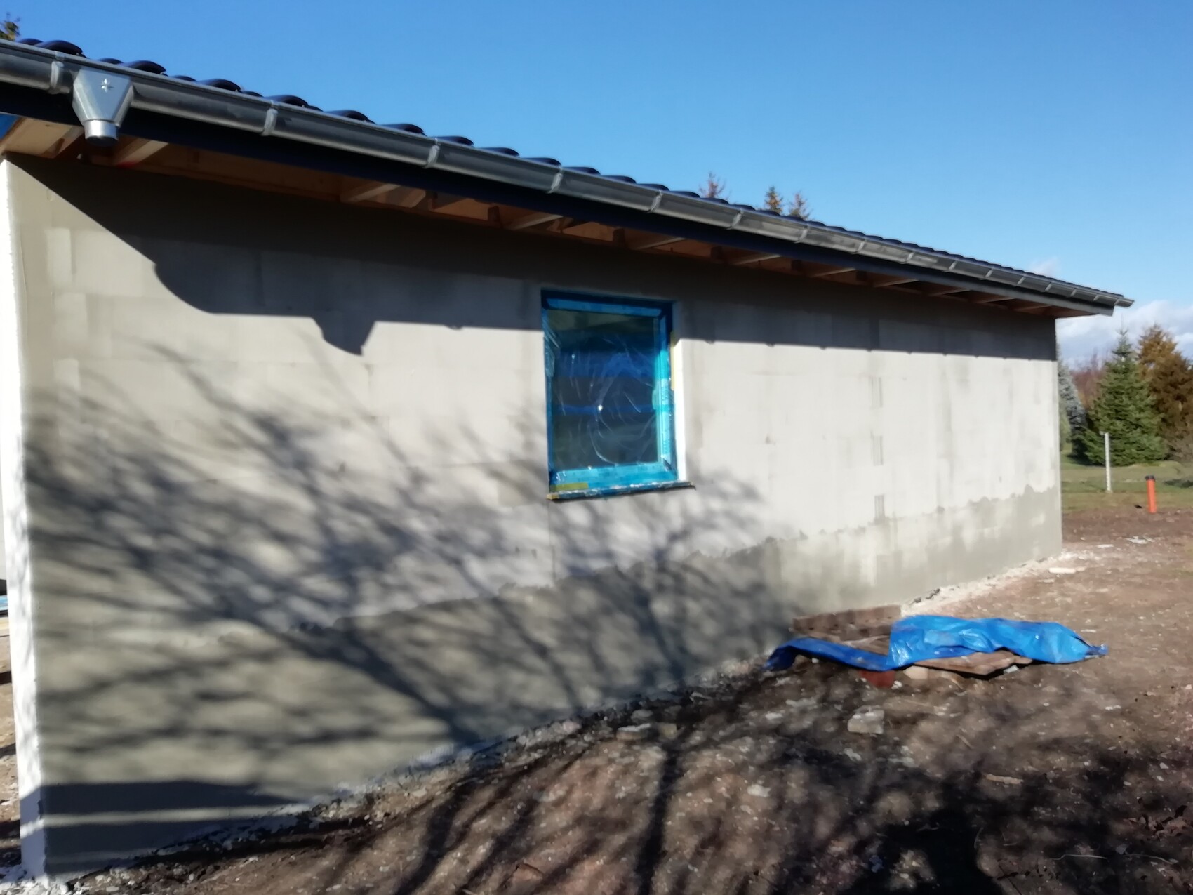 Kleines unfertiges Haus mit grauen Wänden, blau umrahmtem Fenster und Bauabfällen davor.