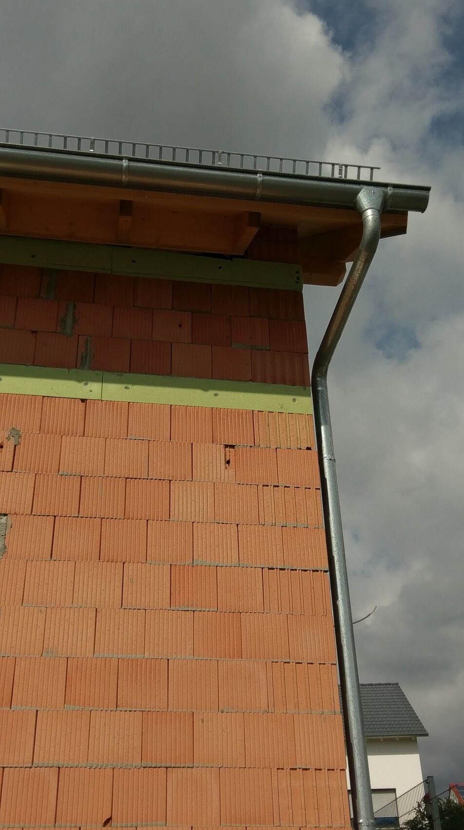Außenansicht eines Rohbaus mit unverputzter Ziegelwand, grünen Dämmstreifen und Dachrinne.