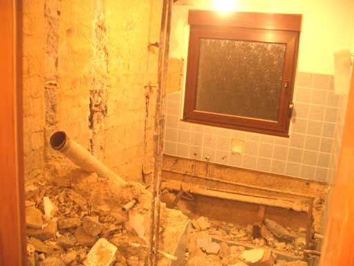 Badezimmer im Rohbau: zerstörte Wand, Schutt, Rohr, Fenster mit Fliesen.