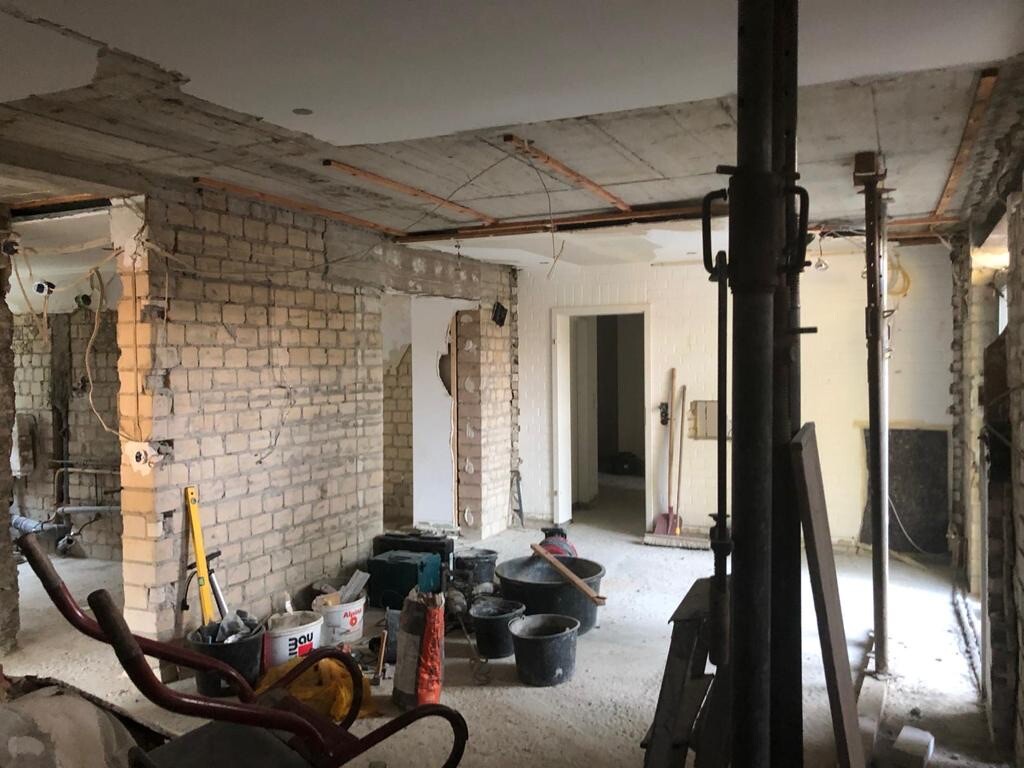Innenraum einer Baustelle: unfertiger Rohbau mit Ziegelwänden, Stützen, Eimern und Werkzeug.