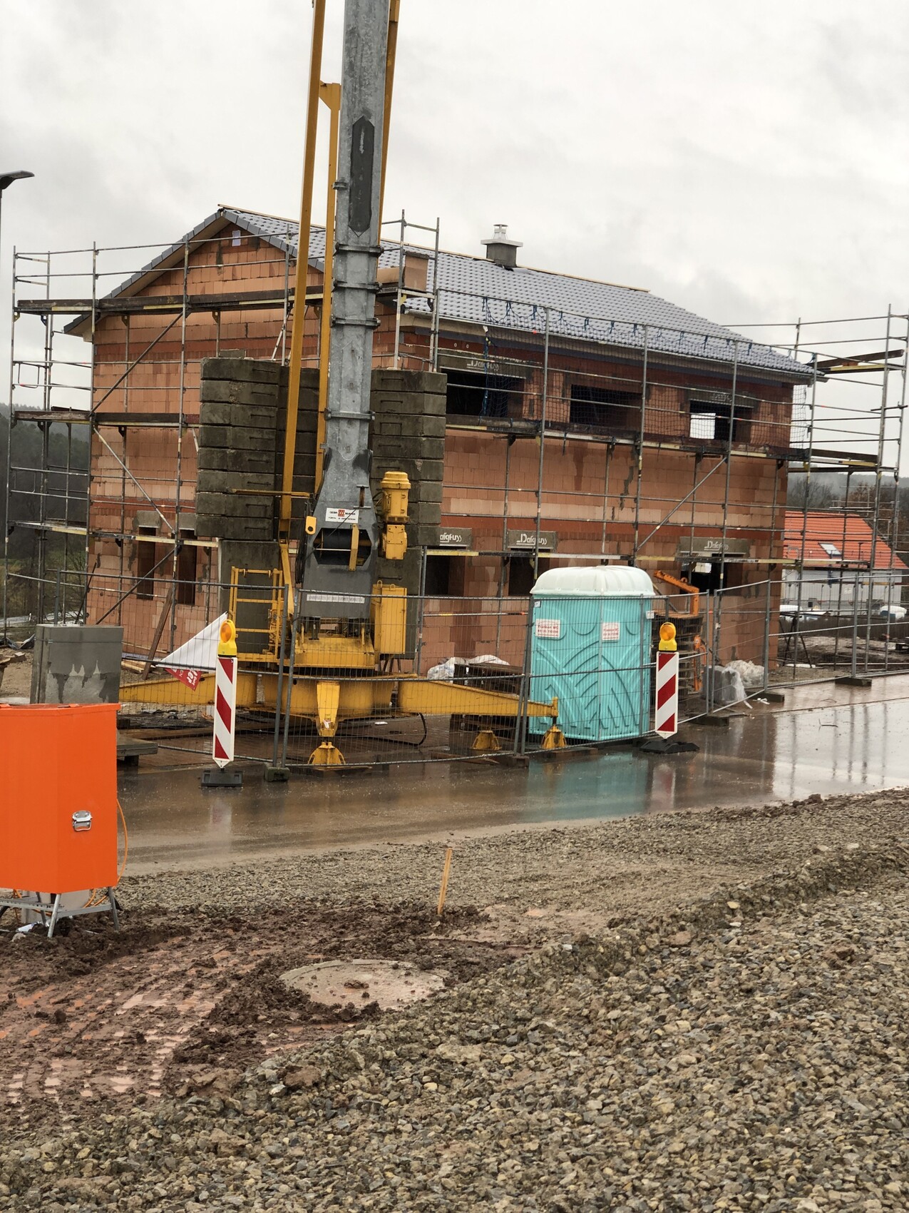 Baustelle: Neubau mit Gerüst, Gelb-Bohrgerät, Porta-Potty und matschigem Gelände.