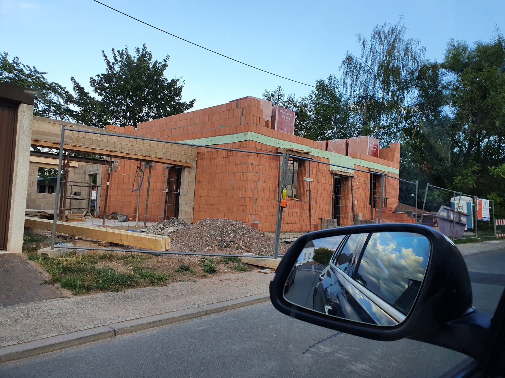 Rohbau eines roten Ziegelgebäudes hinter Bauzaun; Schutt, Gerüst, Straßenszene, Autospiegel.
