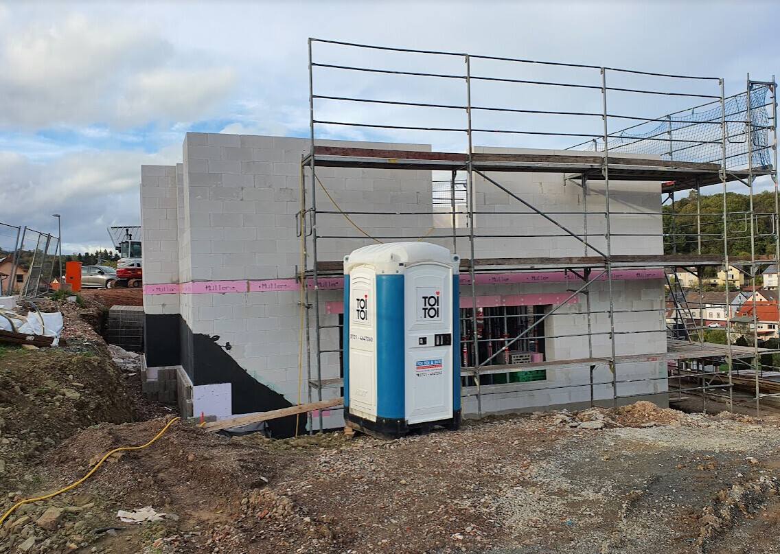 Baustelle: Rohbau eines Hauses mit Gerüst, Betonblöcken und Porta-Toilette.