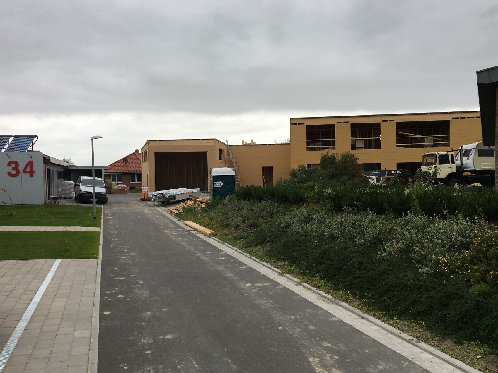 Baugebiet mit unfertigen Gebäuden, Bauholz, Lkw, Container und Grünfläche unter Wolken.