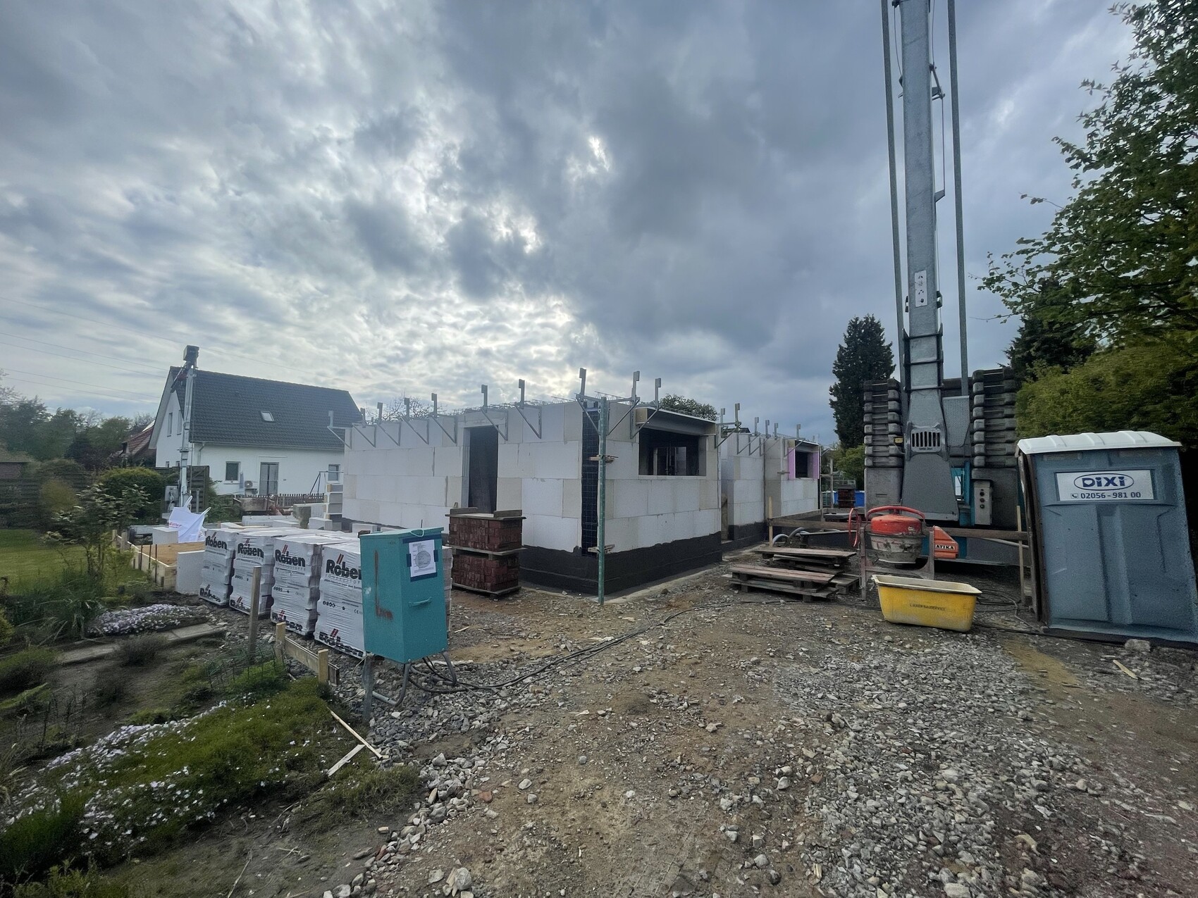 Baustelle mit halbfertigem Haus aus weißen Blöcken, Kran rechts, Baugeräte und Porta-Toilette.