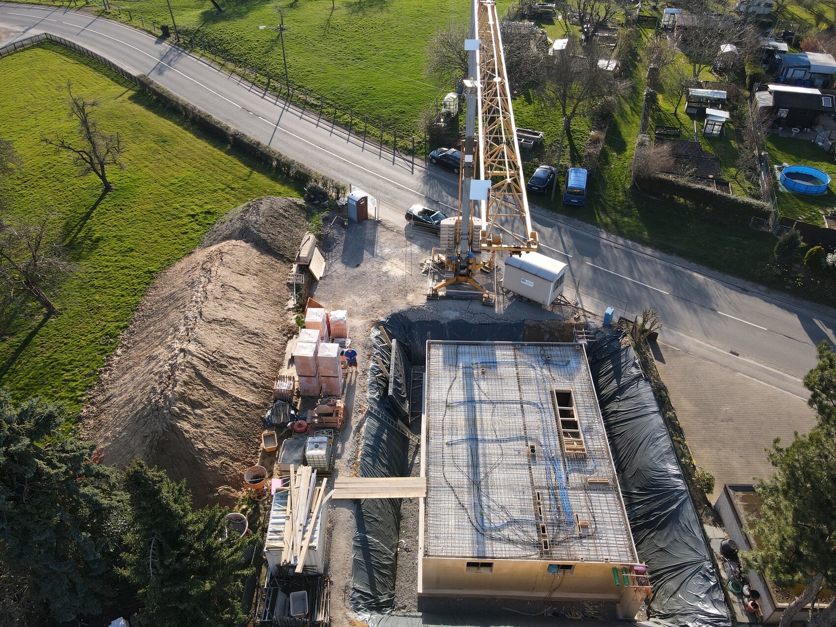 Vogelperspektive: Baustelle mit Kran, Fundament, Sandhügel und Materialstapeln neben Straße.