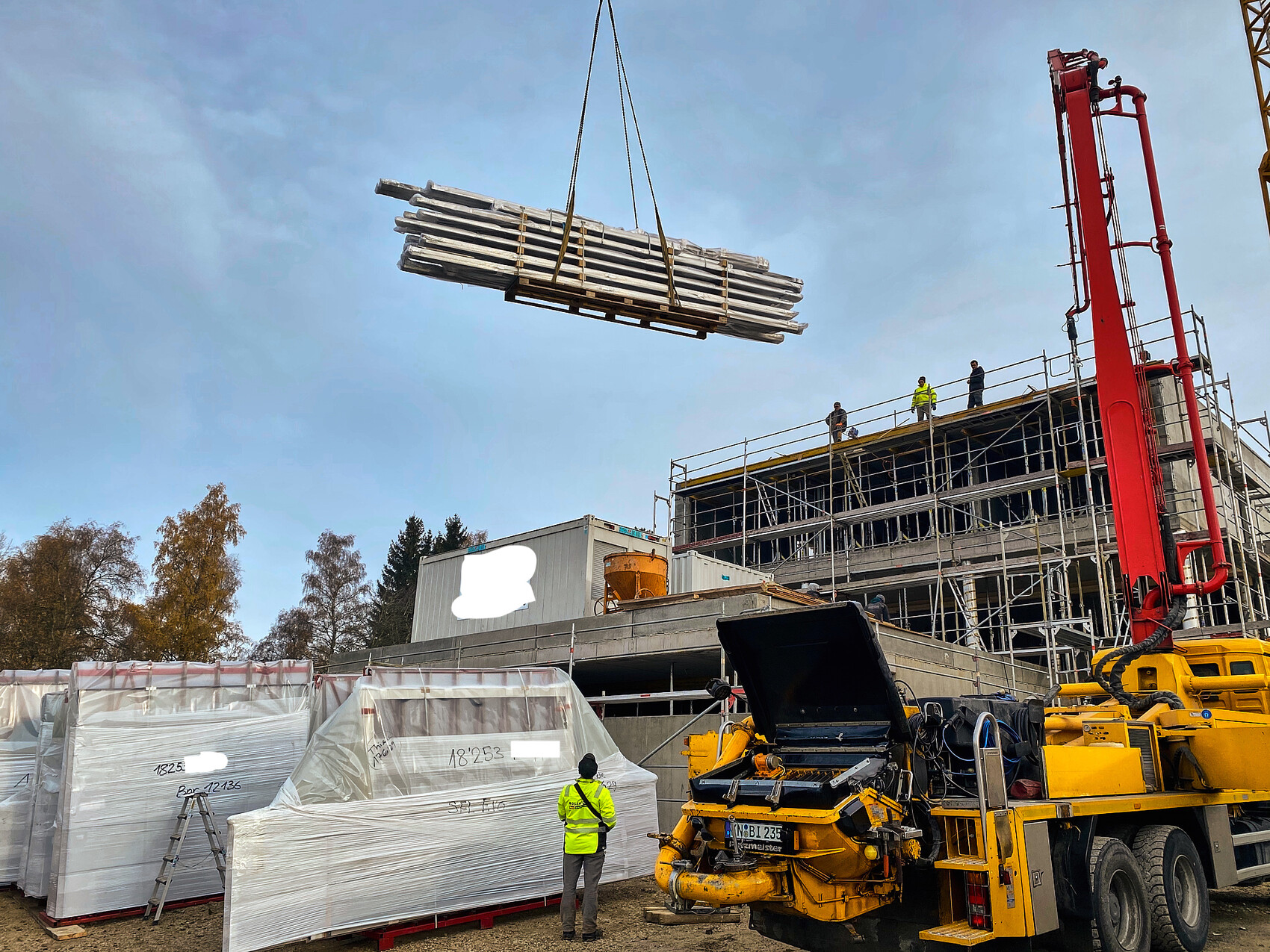 Baustelle: Kran hebt ein Stahlbündel über das Gerüst; Arbeiter beobachten auf dem Bau.
