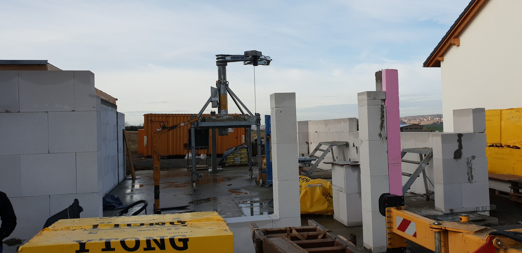 Baustelle mit weißen Betonblöcken, zentralem Hebe-Gerät, gelben Containern und Bauzubehör.