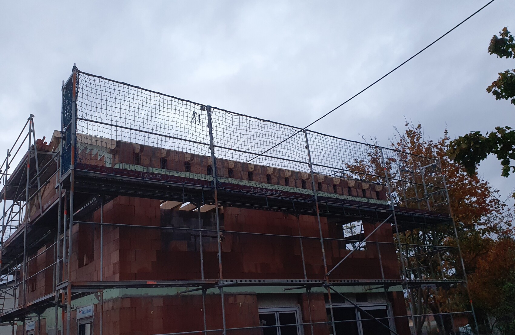 Baugerüst um rotes Backsteinhaus, Netzschutz oben, Herbstbäume daneben.