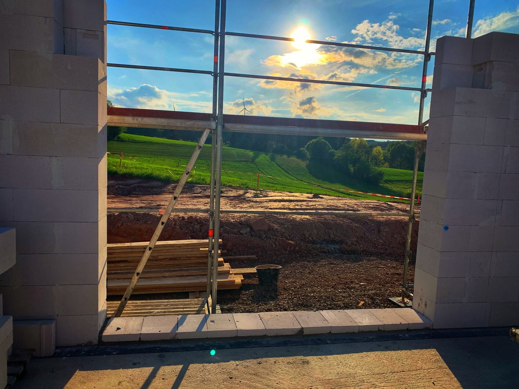Blick durch Baugerüst-Öffnung auf grüne Felder, Bäume, Windrad und sonnigen Himmel.