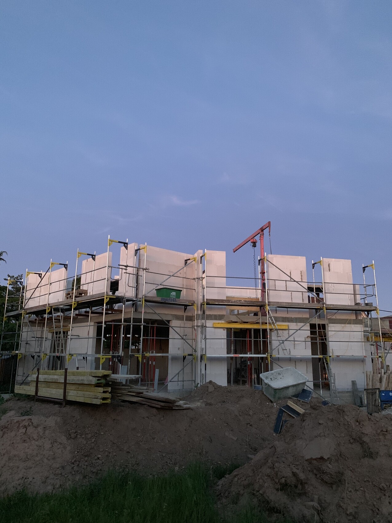 Halbfertiges Gebäude auf Baustelle mit Gerüst, Kran im Hintergrund und Baummaterialien.