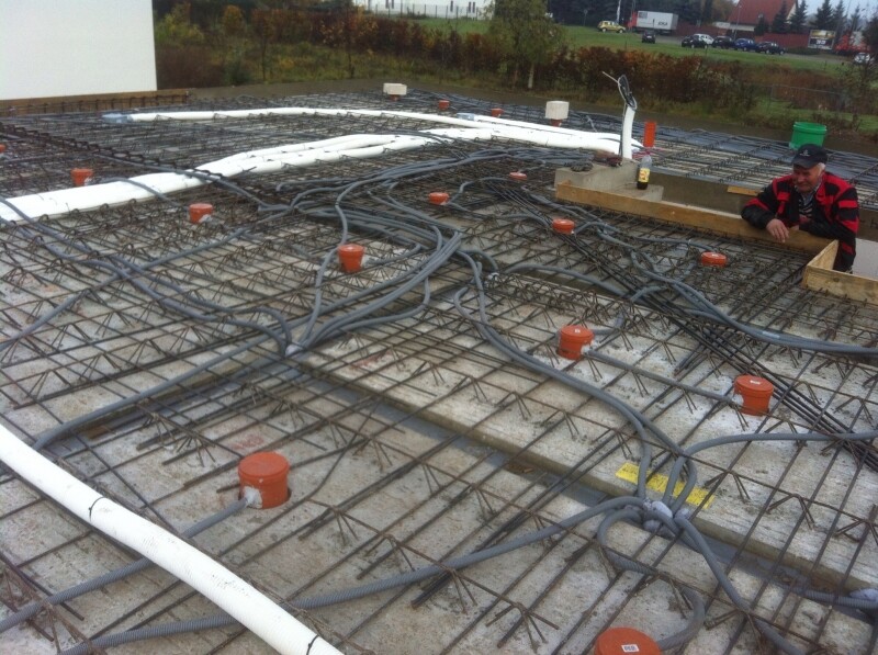 Bewehrungsstahlgitter auf Rohbauboden mit Kabeln und orange Rohrstutzen; Arbeiter am Rand.
