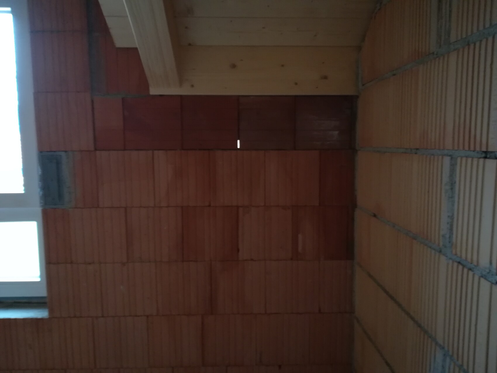 Innenansicht einer Wand aus roten Backsteinen, Holzdecke und Fenster links.