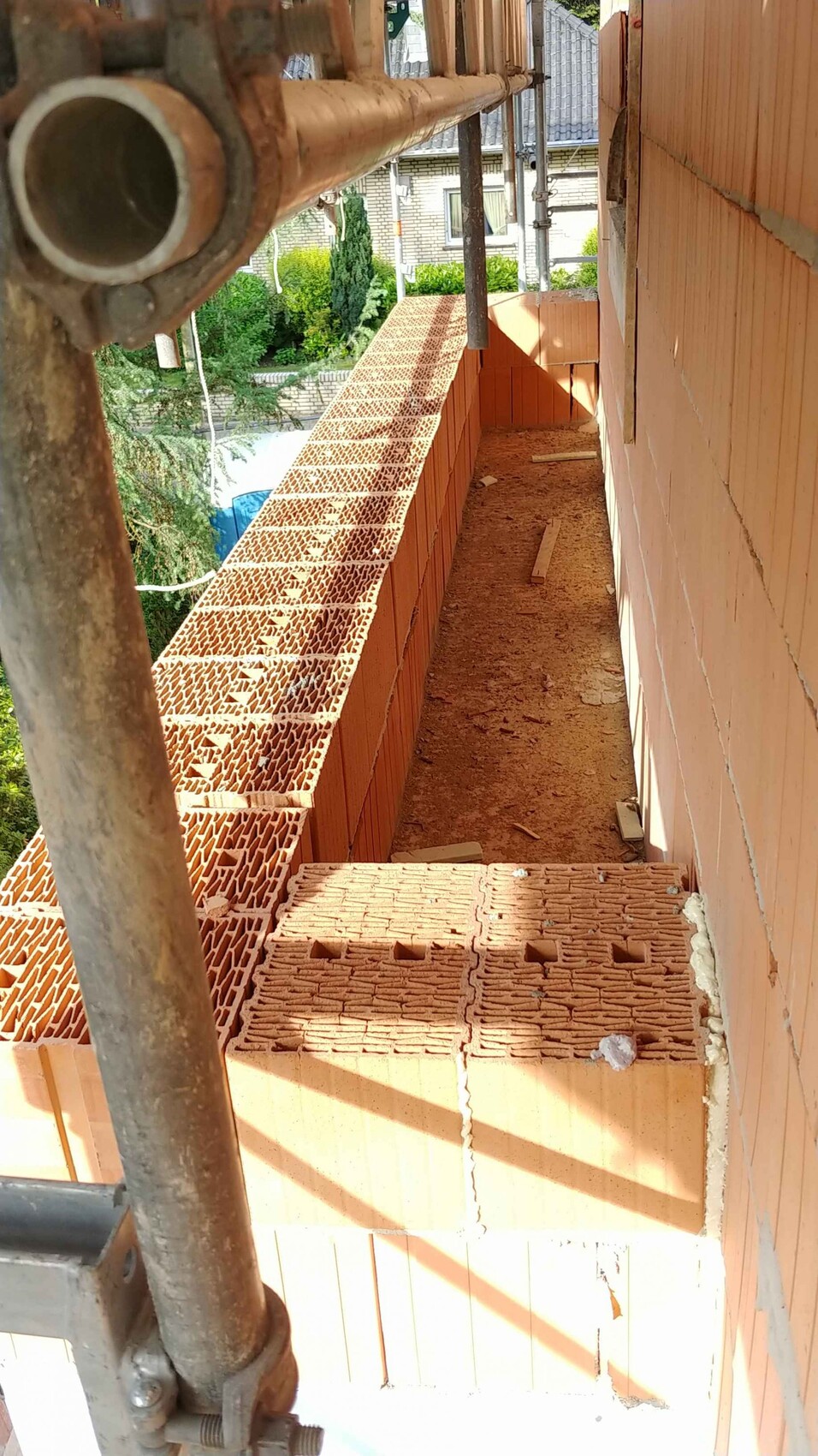Baustelle: orange Ziegelwand, perforierte Ziegel liegen auf dem Gerüst, schmaler Baugang.