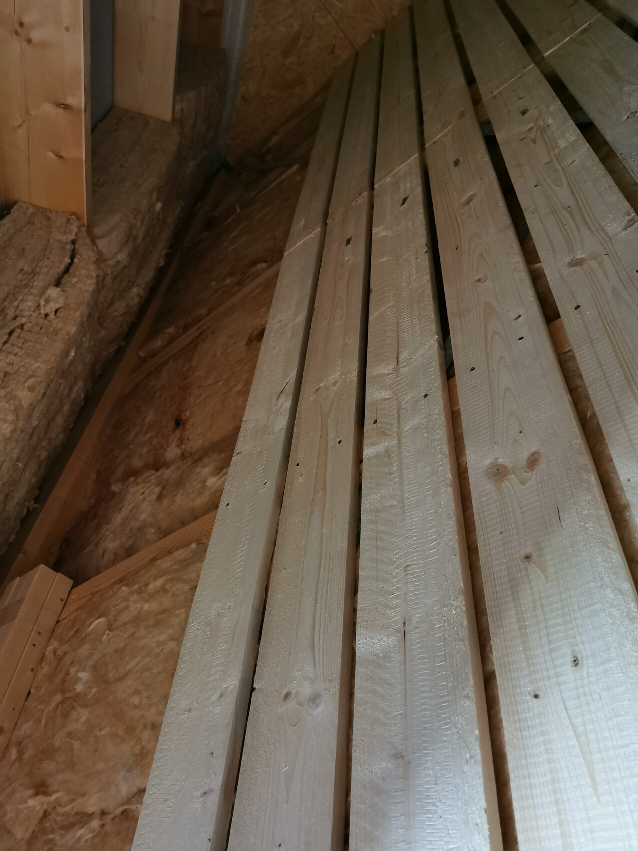 Mehrere lange Holzlatten liegen freiliegend auf einem Dachboden neben Dämmmaterial.