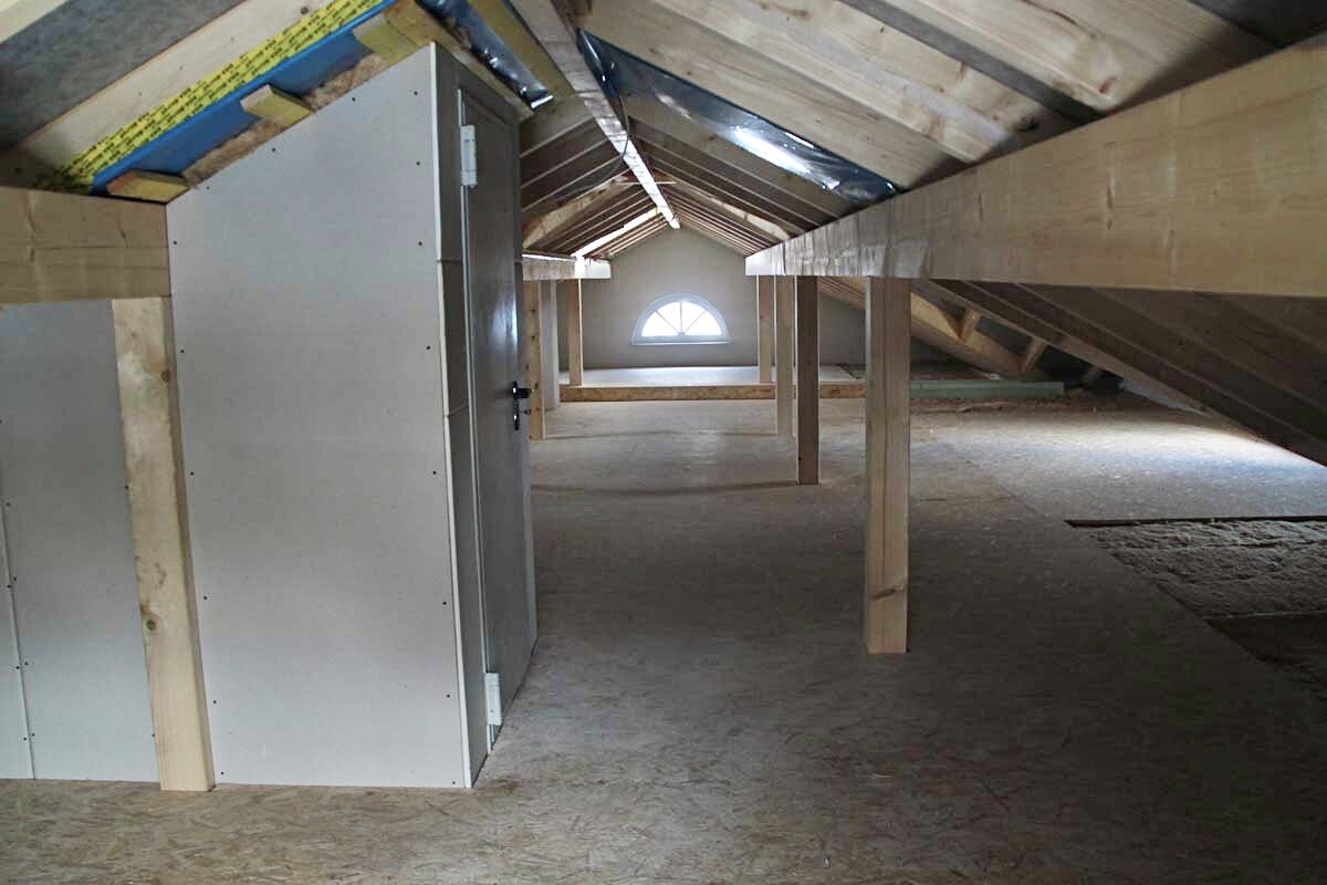Dachboden mit Holzbalken und Stützen; links Metallschrank, hinten halbrundes Fenster.