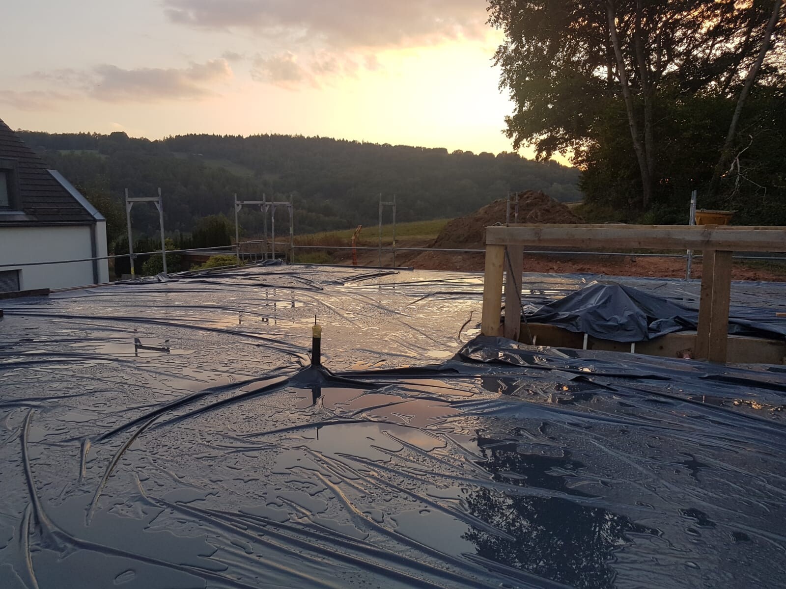 Dachbaustelle mit schwarzer Kunststofffolie, Baugerüst, Erdhügel; Landschaft und Sonnenuntergang