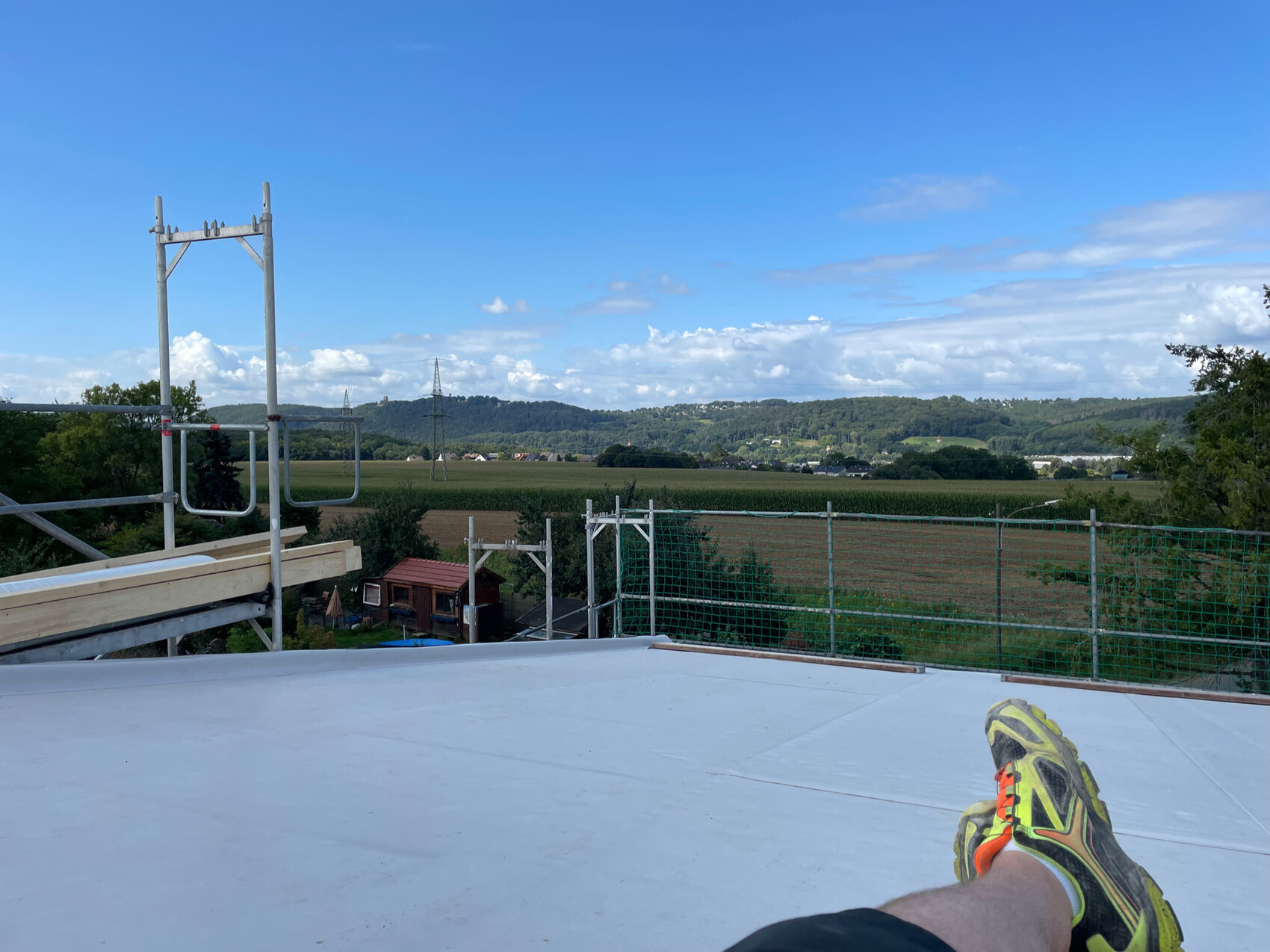 Auf einer Dachfläche sitzende Person mit Sneaker, Baugerüst, Feldlandschaft und Hügel im Hintergrund.