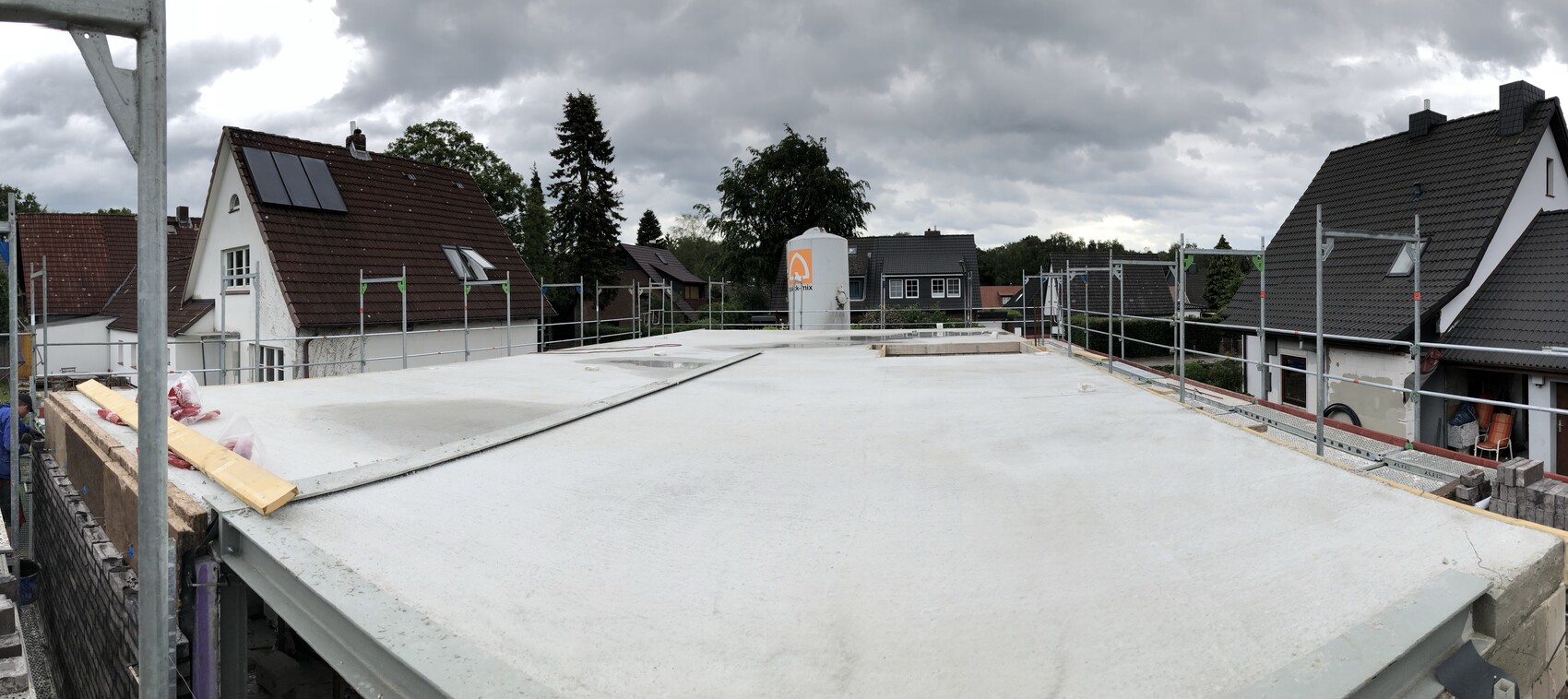 Dachbaustelle mit Betonplatte, umlaufendem Gerüst und Nachbarhäusern unter wolkigem Himmel.