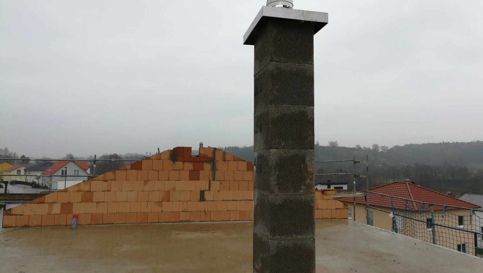 Dachbaustelle: dunkle Stein-Säule mit Metallkappe im Vordergrund, orange Ziegelmauer und Gerüst.
