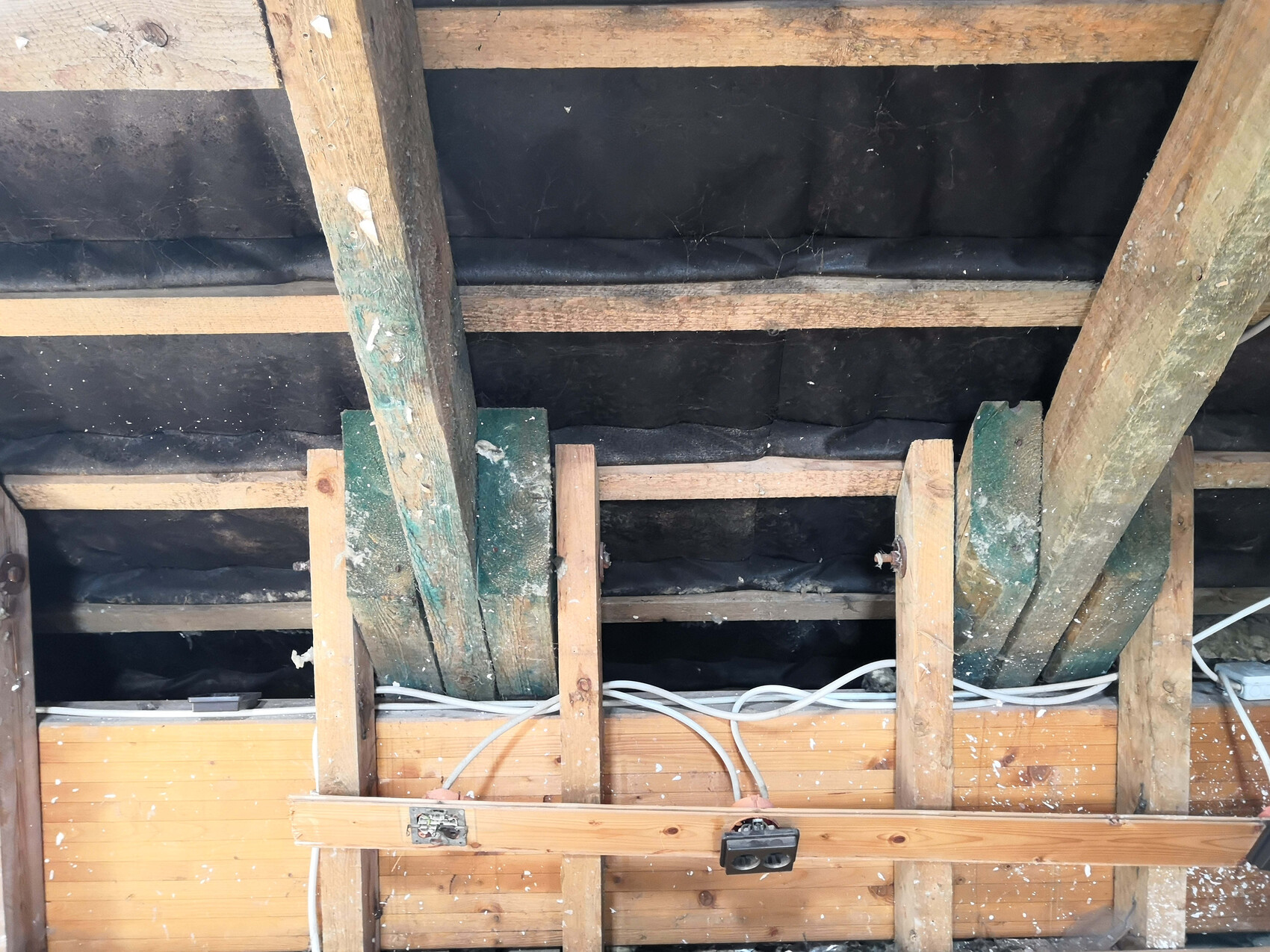 Dachbodenansicht mit Holzbalken, schwarzer Dämmfolie, lose Kabel und einer Steckdose.