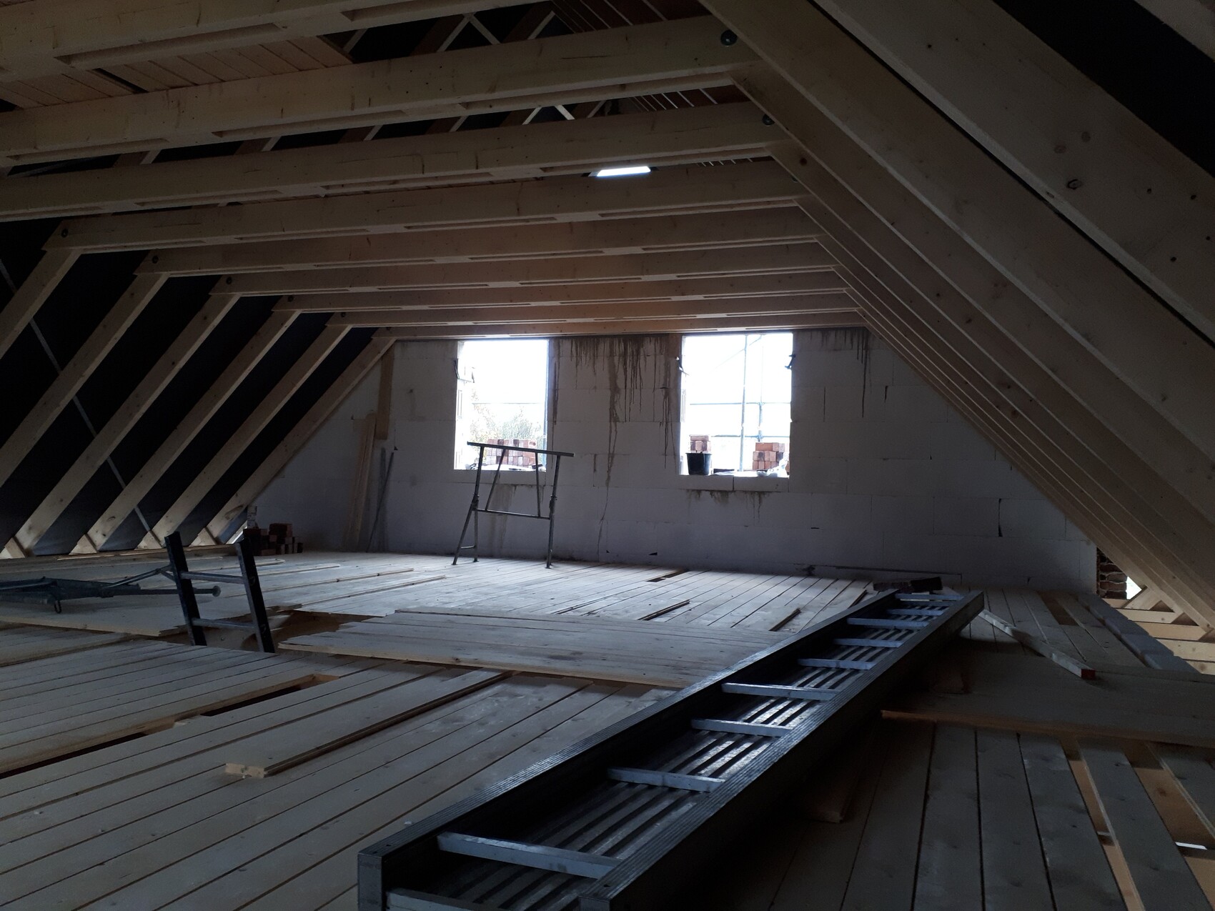 Unfertiger Dachboden mit sichtbarer Holzkonstruktion, Baumaterial, Leiter und zwei Fenster.