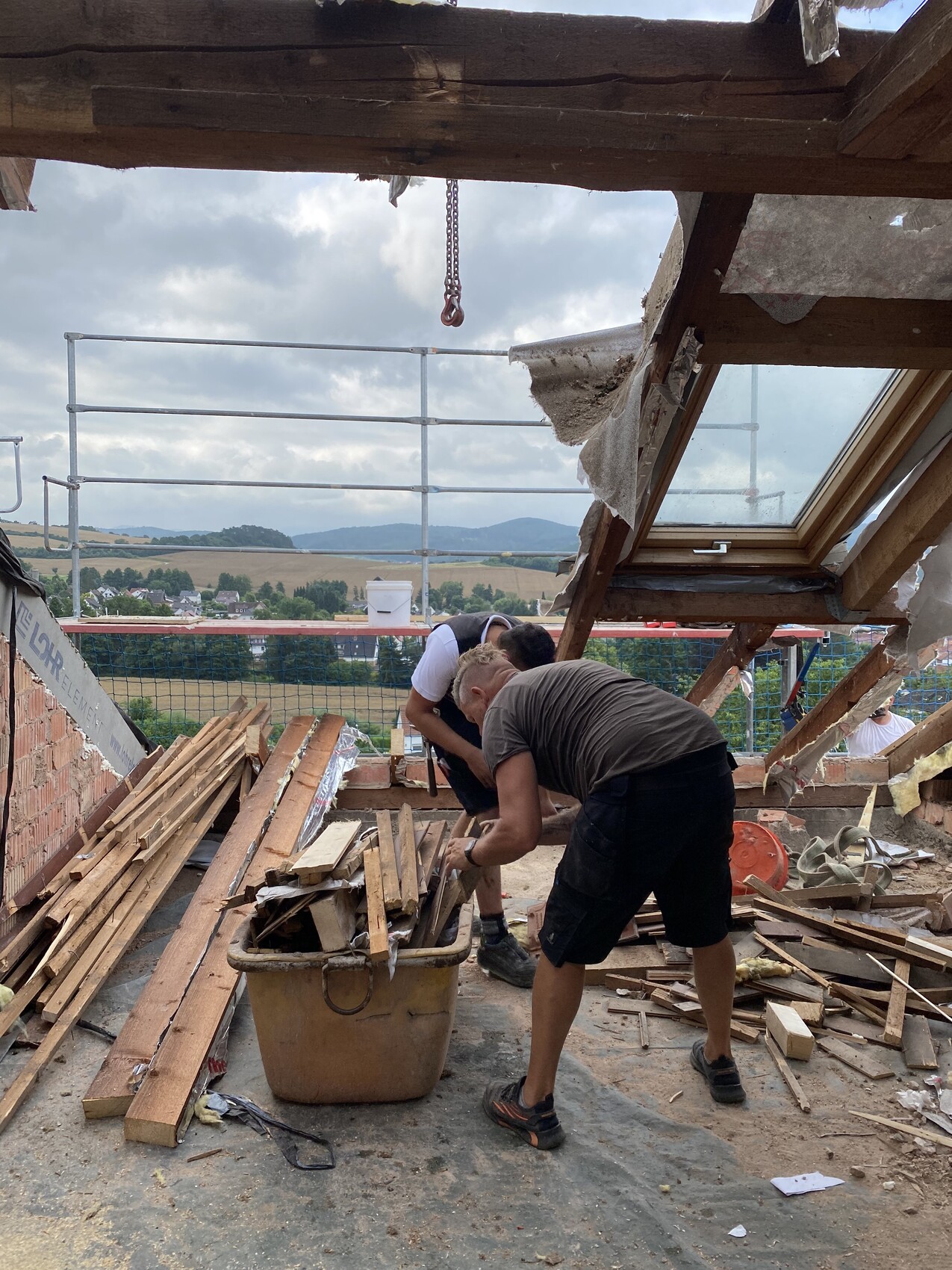 Zwei Arbeiter sortieren Holzreste in einem Behälter auf einer Dachbaustelle, Blick ins Land