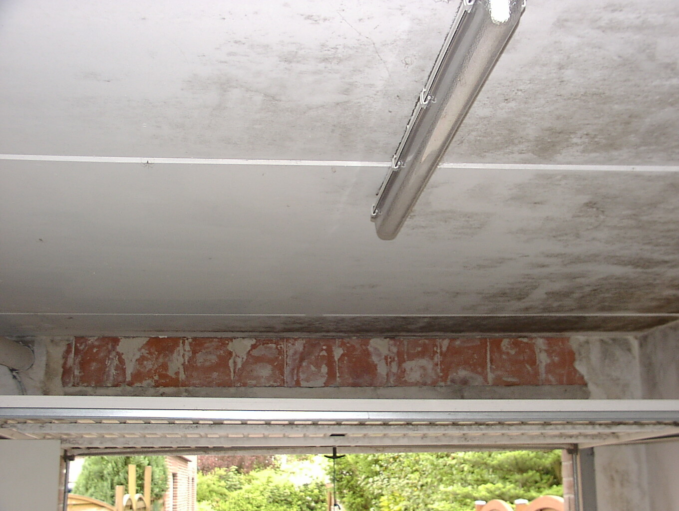 Schräg hängende Leuchte an roher Beton-Decke, Blick aus der Garagenöffnung nach draußen.