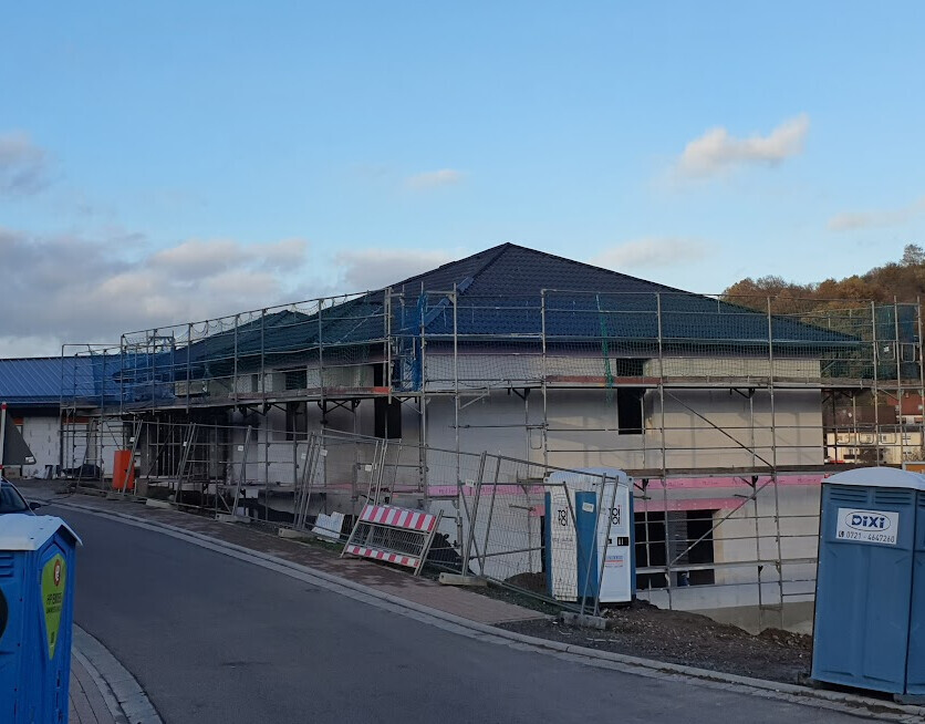 Wohnhaus-Baustelle mit Gerüst, Zaun und Baucontainern am Straßenrand.