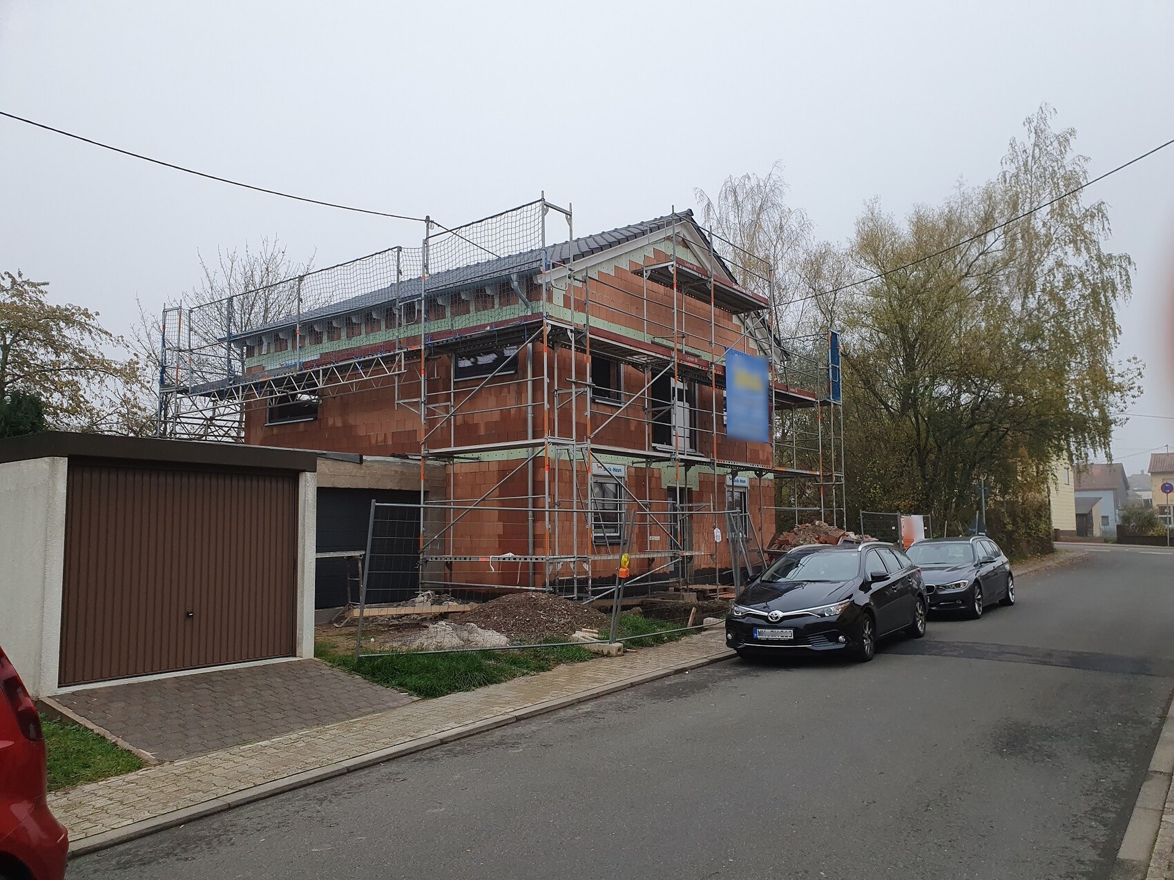 Mehrfamilienhaus im Rohbau mit Gerüst, Bauabfall davor, parkende Autos an der Straße.